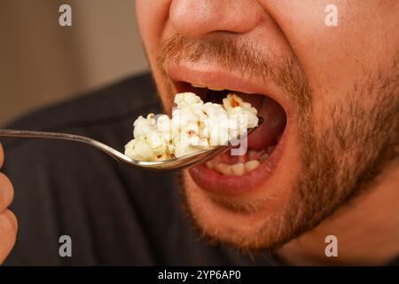 Ein Mann ist dabei, eine Handvoll Popcorn in einem Löffel zu genießen, wobei er sich auf die Details des Imbisses und den Moment der Mahlzeit konzentriert. Ideal für Snacks oder Unterhaltung Stockfoto