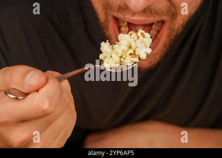 Ein Mann ist dabei, eine Handvoll Popcorn in einem Löffel zu genießen, wobei er sich auf die Details des Imbisses und den Moment der Mahlzeit konzentriert. Ideal für Snacks oder Unterhaltung Stockfoto