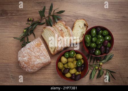 Eine Mischung aus Oliven, Sorten von Chalkidiki, Verdi giganti und Kalamata, in einer Schüssel, mit geschnittenem Ciabata-Brot, Blick von oben, keine Leute Stockfoto