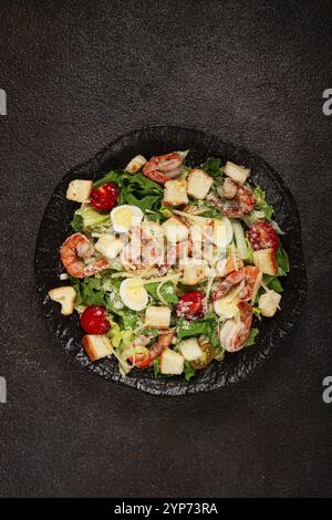 Caesar-Salat mit Tigergarnelen, Garnelensalat, Salatblättern, Wachteleiern, Kirschtomaten, parmesankäse, auf einem schwarzen Teller Stockfoto