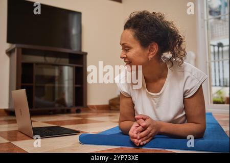 Eine Frau lächelt, während sie an einem Online-Yoga-Kurs teilnimmt. Sie sitzt auf einer blauen Yoga-Matte und beschäftigt sich mit ihrem Laptop. Das gemütliche Zimmer trägt zum entspannten und entspannten Ambiente bei Stockfoto