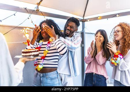 Afrikanische junge Frau mit Augen bedeckt, die eine Geburtstagstorte von multiethnischen Freunden auf der Terrasse empfängt Stockfoto