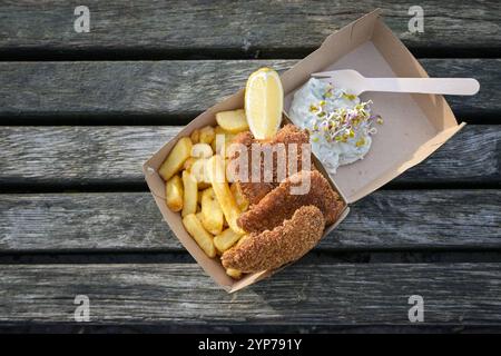 Fish and Chips mit Zitronenscheibe, Tatarsauce und Sprossen in einer Schachtel aus Pappe auf einem verwitterten Holzdock, Fisch und Meeresfrüchte-Snack an der Küste, Kopierraum, Stockfoto