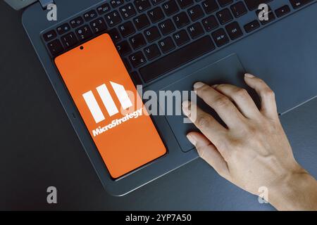 In dieser Fotoabbildung wird das MicroStrategy Incorporated Logo auf einem Smartphone-Bildschirm neben einer Person angezeigt, die ein Notebook verwendet Stockfoto