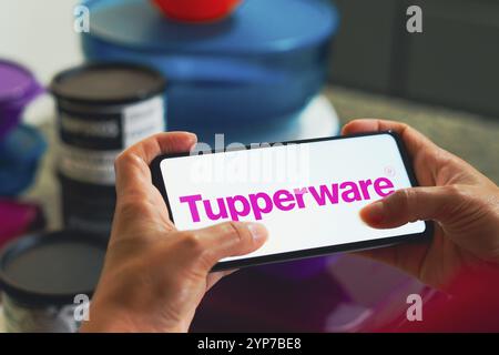 In dieser Fotoabbildung wird das Tupperware-Logo auf einem Smartphone-Bildschirm und im Hintergrund verschiedene Kunststoffprodukte (Kanister) angezeigt. Stockfoto