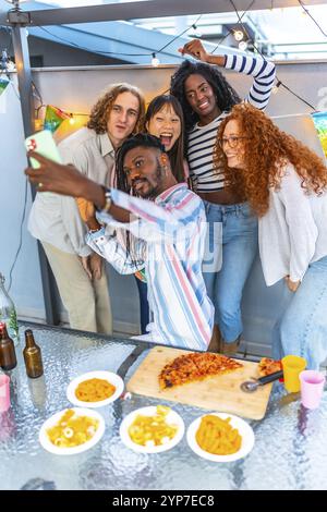 Vertikales Foto eines glücklichen multiethnischen Freundes, der Selfie macht, während er eine Party auf dem Dach feiert Stockfoto