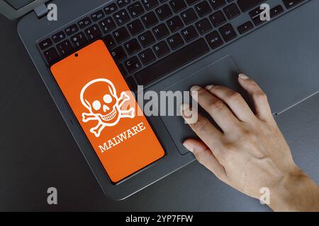 In dieser Fotoabbildung wird das Malware-Symbol auf einem Smartphone neben einer Person angezeigt, die einen Laptop verwendet Stockfoto