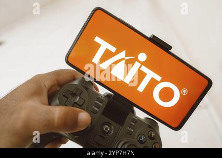 In dieser Fotoabbildung spielt eine Person auf einem Joystick, und das Taito Corporation Logo wird auf einem Smartphone-Bildschirm angezeigt Stockfoto