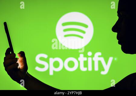 In dieser Fotoabbildung hält die Silhouette einer Frau ein Smartphone mit dem Spotify-Logo im Hintergrund Stockfoto