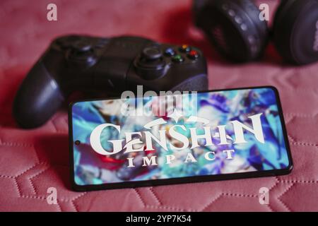 In dieser Fotoabbildung wird das Genshin Impact Logo auf einem Smartphone-Bildschirm neben einem Gamepad angezeigt Stockfoto