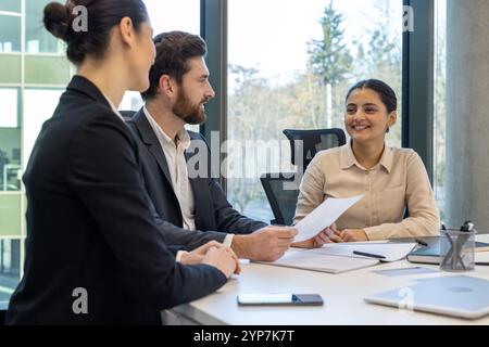 Vorstellungsszene mit einer jungen Frau, die mit zwei Fachleuten in hellem Büroumfeld beschäftigt ist. Die Menschen sitzen mit Dokumenten am Tisch und diskutieren über Möglichkeiten in modernen Arbeitsbereichen. Stockfoto