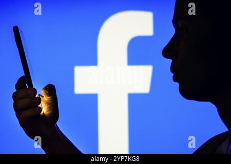 In dieser Fotoabbildung hält die Silhouette einer Frau ein Smartphone mit dem Facebook-Logo im Hintergrund Stockfoto