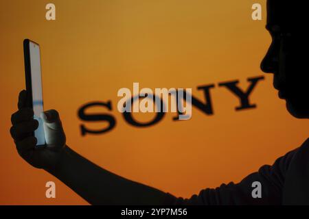 In dieser Fotoabbildung ist das Sony Logo im Hintergrund einer Silhouette einer Frau zu sehen, die ein Mobiltelefon hält Stockfoto