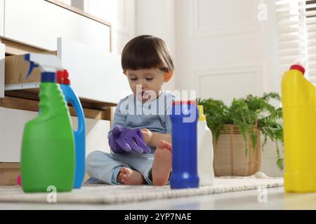 Kleiner Junge, der mit Handschuhen zwischen Waschmittelflaschen in der Nähe des Schranks zu Hause spielt. Kind in Gefahr Stockfoto