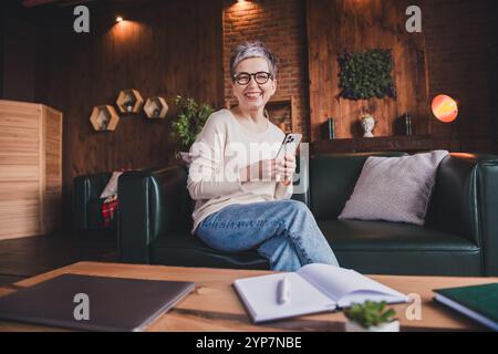 Charmante ältere Frau mit kurzen grauen Haaren und Pullover sitzt bequem auf einer Couch in einem modernen Innenraum Stockfoto