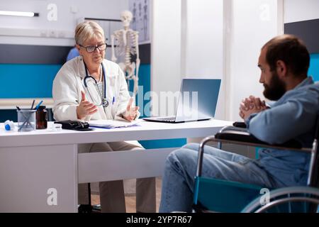 Kaukasischer männlicher Rollstuhlfahrer hört aufmerksam auf ältere Ärztinnen, die seine medizinische Behandlung am Kliniktisch diskutieren. Oberarzt, der mit bärtigem Mann über seine Gesundheitsziele spricht. Stockfoto