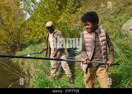 Mittlere Nahaufnahme eines aufgeregten Jungen mit einer riesigen Angelrute in seinen kleinen Händen, der versucht, den ersten Fisch zu fangen Stockfoto