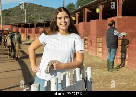 Glückliche junge Frau in lässiger Kleidung steht neben dem Zaun und lächelt vor der Kamera während des Besuchs der Pferdefarm Stockfoto