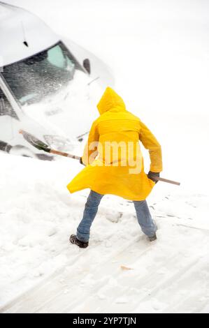 Person in gelbem Regenmantel, die bei starkem Schneefall Schnee von einem Auto schaufelt, Stockfoto