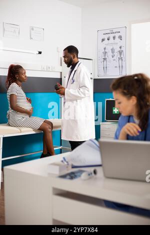 Im Hintergrund spricht Arzt mit Flasche Pillen zu schwangeren schwarzen Frauen, während junge weibliche Krankenschwester medizinische Aufzeichnungen liest. Der männliche Arzt gibt der werdenden Mutter verordnete Ergänzungen. Stockfoto