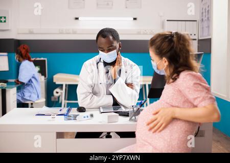 Die erwartende Mutter sitzt am Schreibtisch und berät sich mit ihrem Arzt, während die Krankenschwester im Hintergrund einen Desktop-pc verwendet. Schwangere weiße Frau und männlicher Geburtshelfer mit Gesichtsmasken, die Gesundheitsdokumente überprüfen. Stockfoto