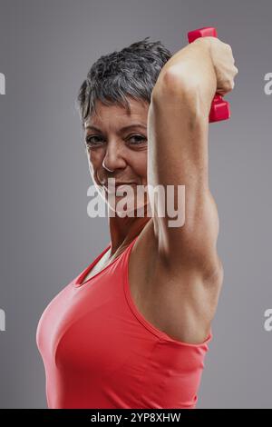 Eine entschlossene Seniorin mit rotem Tanktop hebt eine rosa Kurzhantel und lächelt selbstbewusst in die Kamera und zeigt ihre Stärke und ihr Engagement Stockfoto