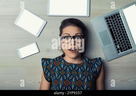 Die junge Erwachsene Frau, die mit mehreren elektronischen Geräten auf dem Boden liegt, fühlt sich von der Überladung von Technologie und Informationen überwältigt Stockfoto