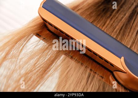 Kupferfarbener und dunkelblauer Haarglätter gleiten durch langes, hellblondes Haar. Kosmetikmode und Styling-Werkzeuge Stockfoto