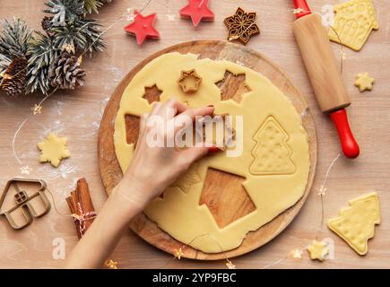 Frau, die weihnachtskekse mit unterschiedlich geformten Keksschneidern auf einem Holzbrett zubereitete, umgeben von festlichen Dekorationen Stockfoto