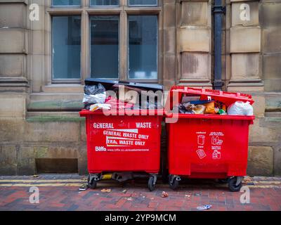 2 hellrote Biffa-Mülltonnen, die vor einem alten Steingebäude mit Müll überlaufen. Stockfoto