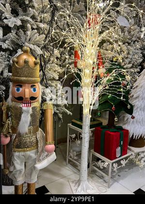 Dieses vertikale Foto zeigt eine königliche Nussknacker-Figur, die mit goldenen Details verziert ist und neben einer leuchtenden, baumähnlichen Dekoration steht, die in warmen Farben gewickelt ist Stockfoto