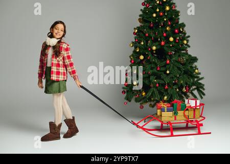 Ein glückliches Mädchen in kuscheliger Kleidung zieht einen bunten Schlitten voller Geschenke an einen Weihnachtsbaum. Stockfoto