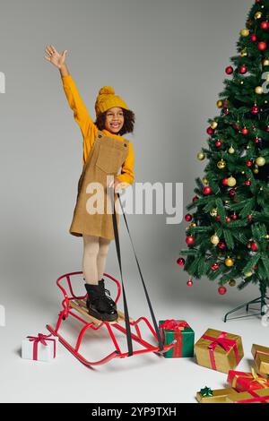 Das Mädchen steht fröhlich auf einem Schlitten, umgeben von Geschenken und einem festlichen Weihnachtsbaum. Stockfoto