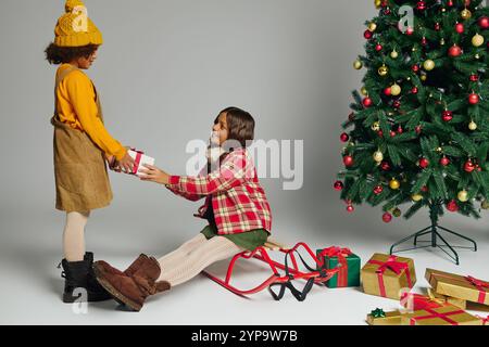 Zwei junge Mädchen teilen sich einen wunderbaren Moment, indem sie Geschenke an einem wunderschön geschmückten Baum austauschen. Stockfoto