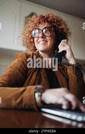 Rothaarige Frau mittleren Alters, die Laptop benutzt und telefoniert. Stockfoto