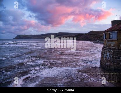 Ein kalter Wintertag endet mit einem pulsierenden Sonnenuntergang, während stürmische Winde starke Wellen in der Robin Hoods Bay hinauf treiben. Stockfoto