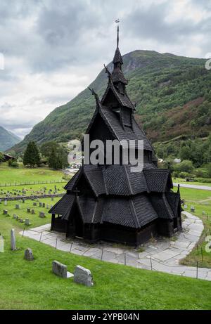 Borgund Stave Church in Norwegen, ein schwarzer hölzerner Wikingertempel, umgeben von Grabsteinen und im Hintergrund ein Berg unter bewölktem Himmel. Norw Stockfoto