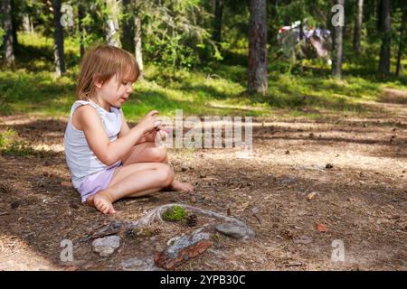 Ein kleines Mädchen in grauem Tanktop und hellvioletten Shorts sitzt barfuß auf dem Waldboden und spielt mit kleinen Gegenständen in ihren Händen. Die Sonnenfilter t Stockfoto