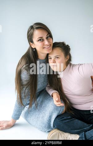 Eine Mutter und ihre Tochter mit Down-Syndrom sitzen zusammen und lächeln. Die Mutter hat ihren Arm um ihre Tochter und sie sehen beide aus Stockfoto
