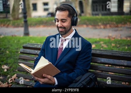 Ein junger Geschäftsmann in Anzug und Krawatte sitzt auf der Bank im Park, liest Bücher und hört Musik mit Kopfhörern und genießt seine Freizeit Stockfoto