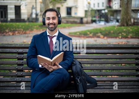 Ein lächelnder Geschäftsmann mit Anzug und Kopfhörern sitzt auf einer Bank im Park, hört ein Hörbuch und hält ein Buch in der Hand Stockfoto