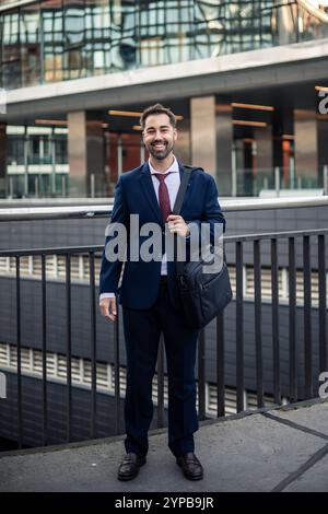 Porträt eines selbstbewussten Geschäftsmannes, der lächelnd auf einer Brücke vor modernen Bürogebäuden steht, eine Laptoptasche trägt und Professi ausstrahlt Stockfoto