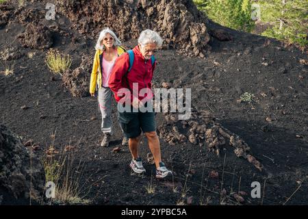 Aktives Seniorenpaar mit farbenfrohen Jacken, das vulkanisches Gelände erkundet. Natürlicher ehrlicher Moment abenteuerlicher Rentner, die auf einem dramatischen Lavafeld wandern, EM Stockfoto