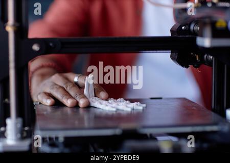 Nahaufnahme einer Person, die einen 3D-Drucker bedient, mit detaillierter Ansicht der Hand und des gedruckten Objekts. Roter Pullover im Hintergrund, während der Fokus auf den 3D-Druckprozess gerichtet bleibt Stockfoto