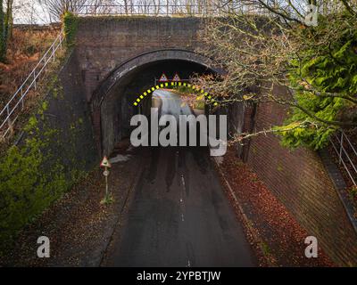 Nasse Asphaltstraße mit Warnschildern, die während der Herbstsaison unter einer niedrigen Ziegelbrücke in einem ländlichen Gebiet vorbeiführen Stockfoto