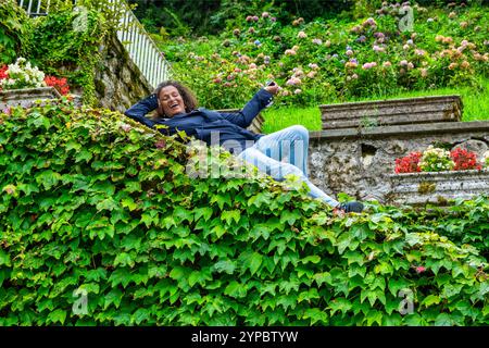 Amüsante, schöne südamerikanische Frau, mittleres Alter mit lockigen Haaren, zwischen Blättern und Blumen gelegen. Stockfoto