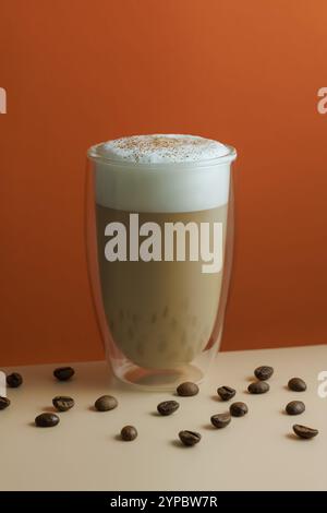 Ein Glas Cappuccino und Kaffeebohnen in der Nähe des Tisches auf dem orangefarbenen Hintergrund Stockfoto