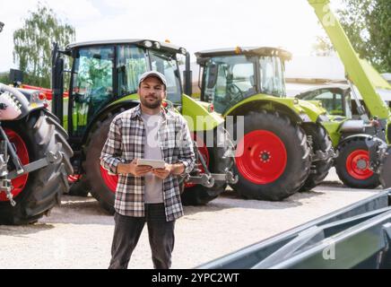 Kaukasischer Mann in kariertem Hemd, der mit einem digitalen Tablet neben Traktoren in einem Landmaschinenhändler steht. Stockfoto