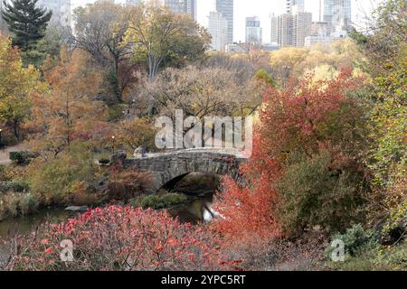 Herbstlaub schmücken die Landschaft rund um eine Steinbrücke im Central Park und vermischen leuchtende Rot-, Gelb- und Orangen vor dem Hintergrund von Gebäuden Stockfoto