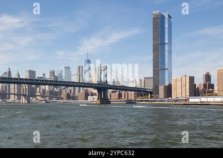 Die Manhattan Bridge überquert den Fluss und verbindet Brooklyn mit Manhattan. Wolkenkratzer erheben sich unter blauem Himmel über der Skyline. Stockfoto